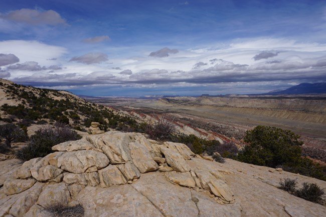 View from atop a sandstone rim into a long valley with mesas and tall mountains on the opposite side