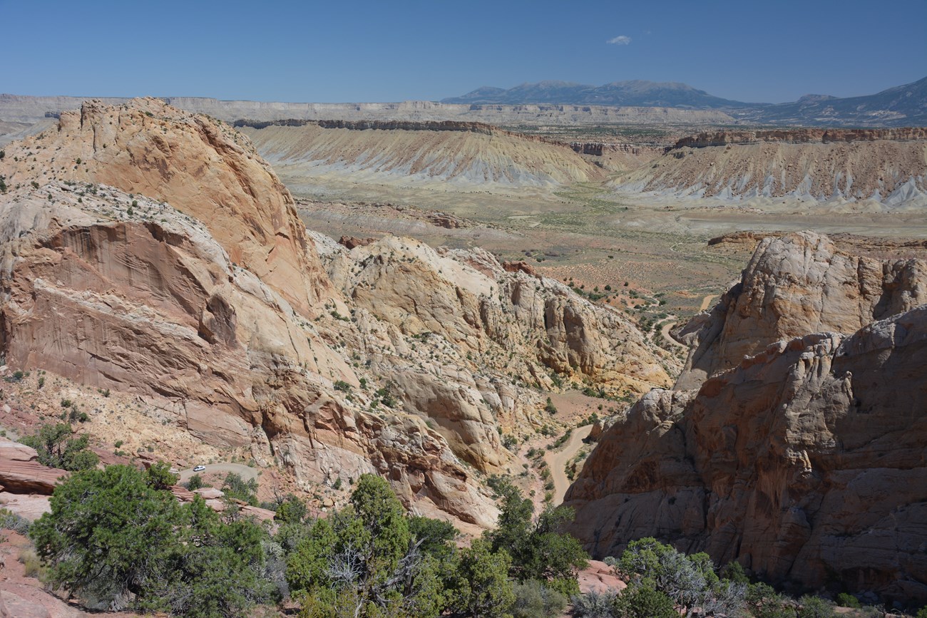 Panoramic view of rock cliffs, some trees, a dirt road, and mountains in the distance, with blue sky.
