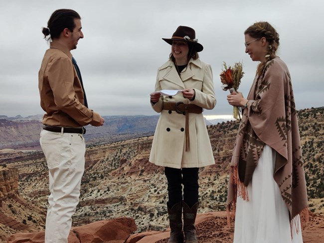 A man with hair in a bun, dress shirt, and tie smiling at a woman across from him who is holding a bouquet of dried flowers, and wearing a white dress and shawl, while another woman in a white jacket holds a piece of paper, in front of a canyon.