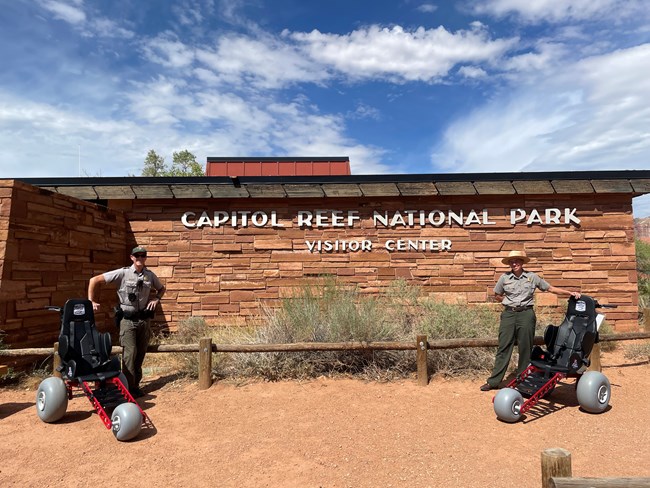 Two NPS rangers stand beside two all-terrain wheelchairs in front of the Capitol Reef National Park Visitor Center sign.