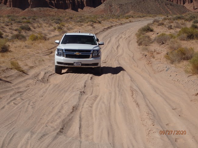 A white SUV drives over deep sand ruts on an unpaved road.