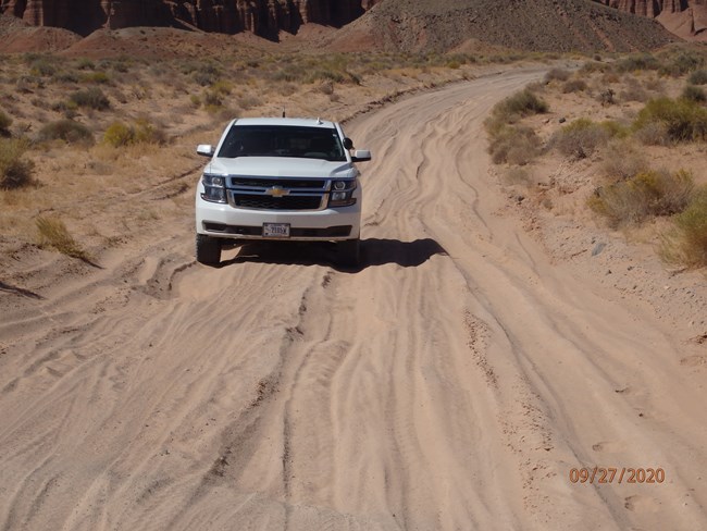 A white SUV drives over deep sand ruts on an unpaved road.