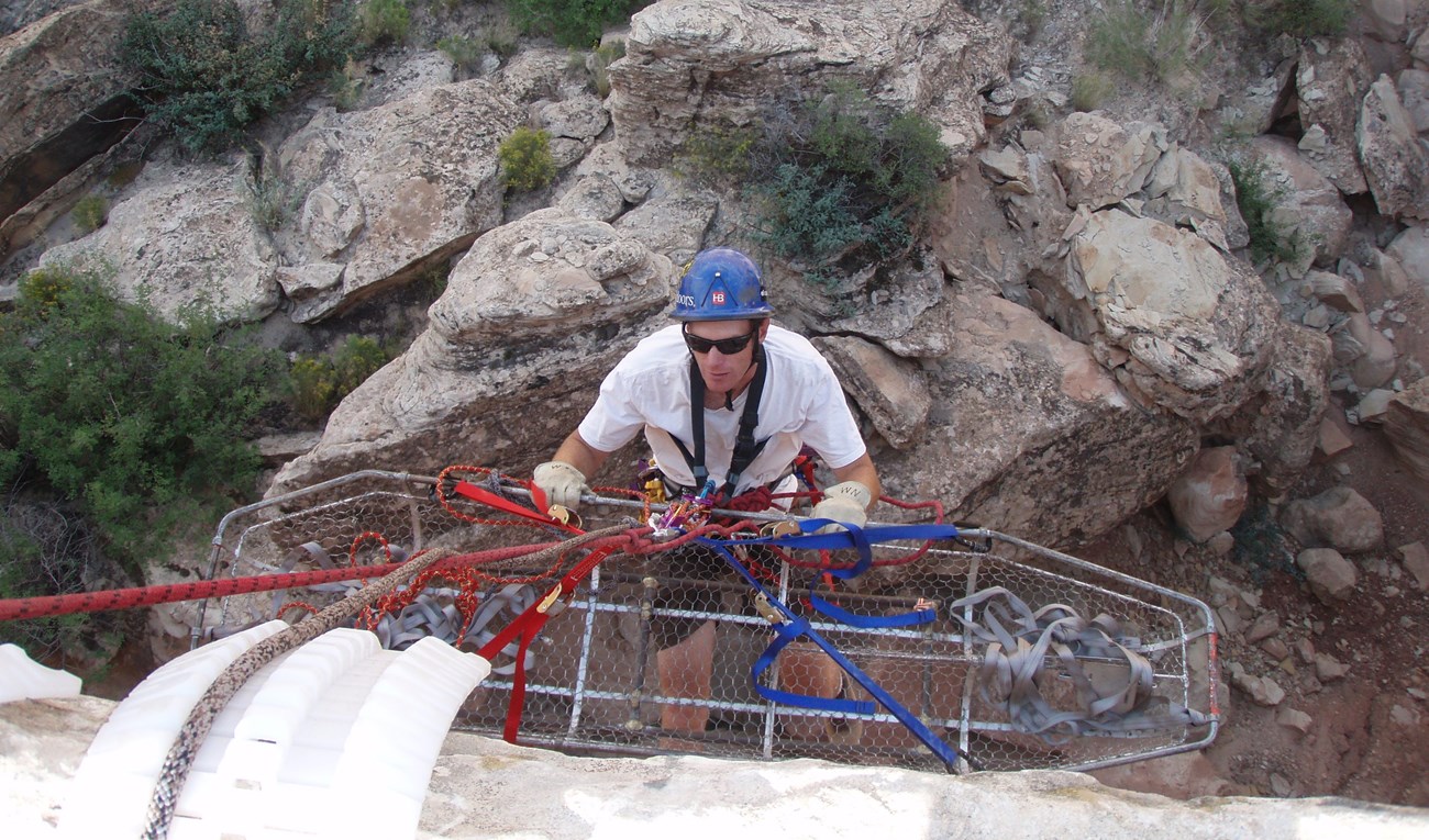 A rappeller with hard hat moves with a metal basket for patients down a cliff.