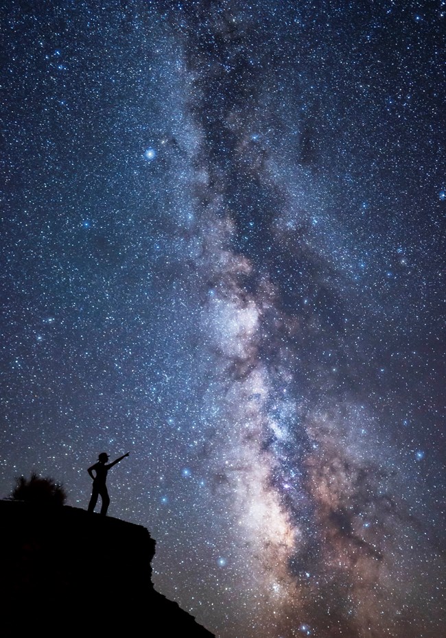 A sky full of stars with the denser milky way in the middle. A person in silhouette points to the stars.