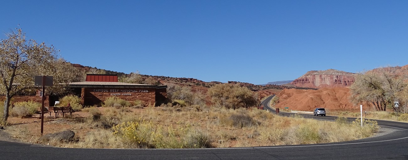 Low-profile stone building that blends into the redrock landscape, with blacktop highway and road to the right and in front of it. Blue sky above.