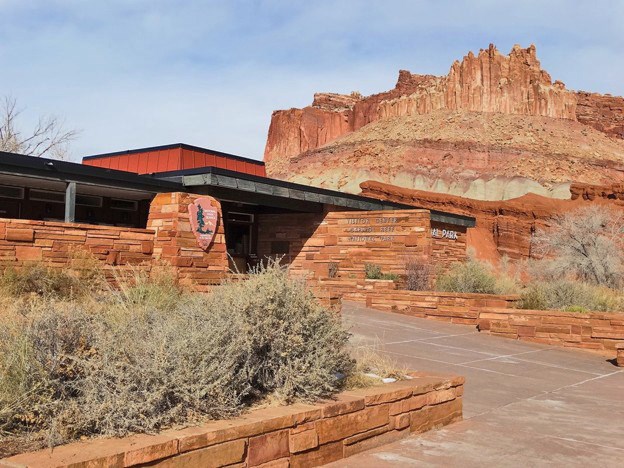 Capitol Reef visitor center, a low red sandstone building with the NPS arrowhead on its front wall, below a towering red rock formation
