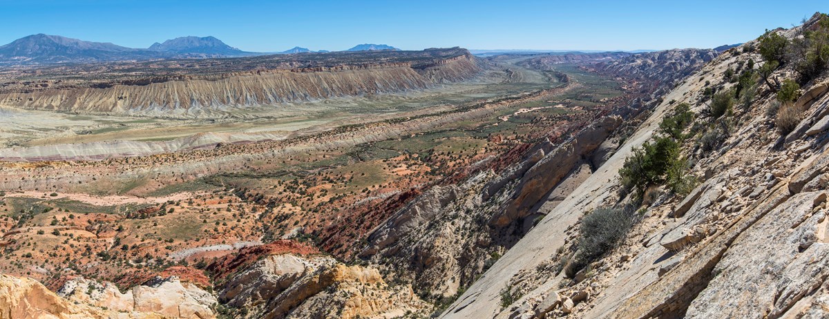 Long, wide valley with ridges running parallel inside it, with many colorful rock layers and blue sky above.