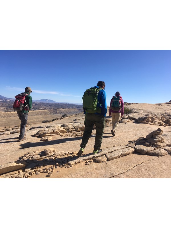 Three people walking on tan sandstone, with some sparse vegetation, and views into a wide valley and blue sky above.