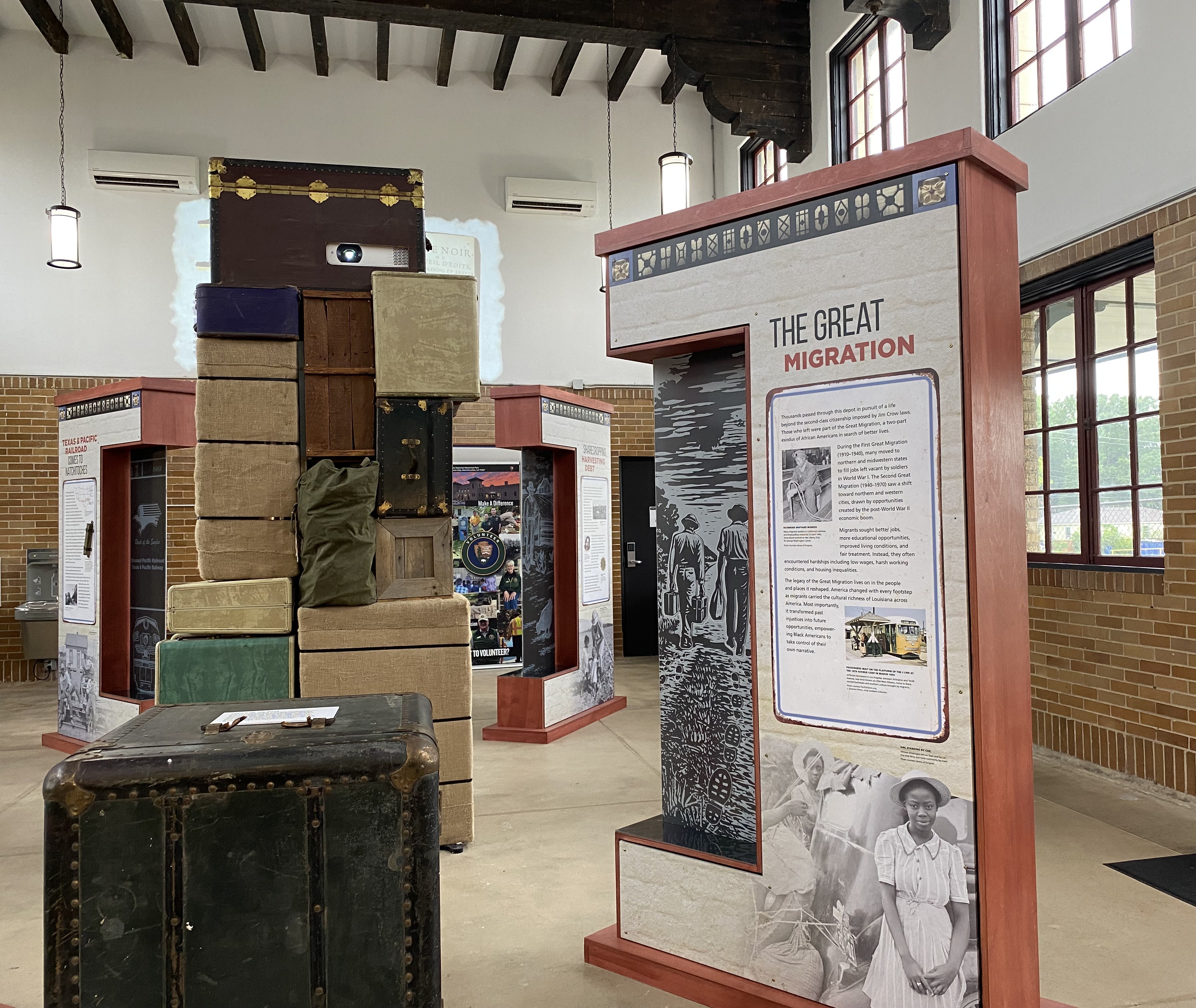 Interior view of the Texas & Pacific Railway Depot museum at Cane River Creole National Historical Park. In the center of the exhibit hall, a tall sculpture made from stacked vintage trunks, suitcases, wooden crates, and travel bags symbolizes the journey