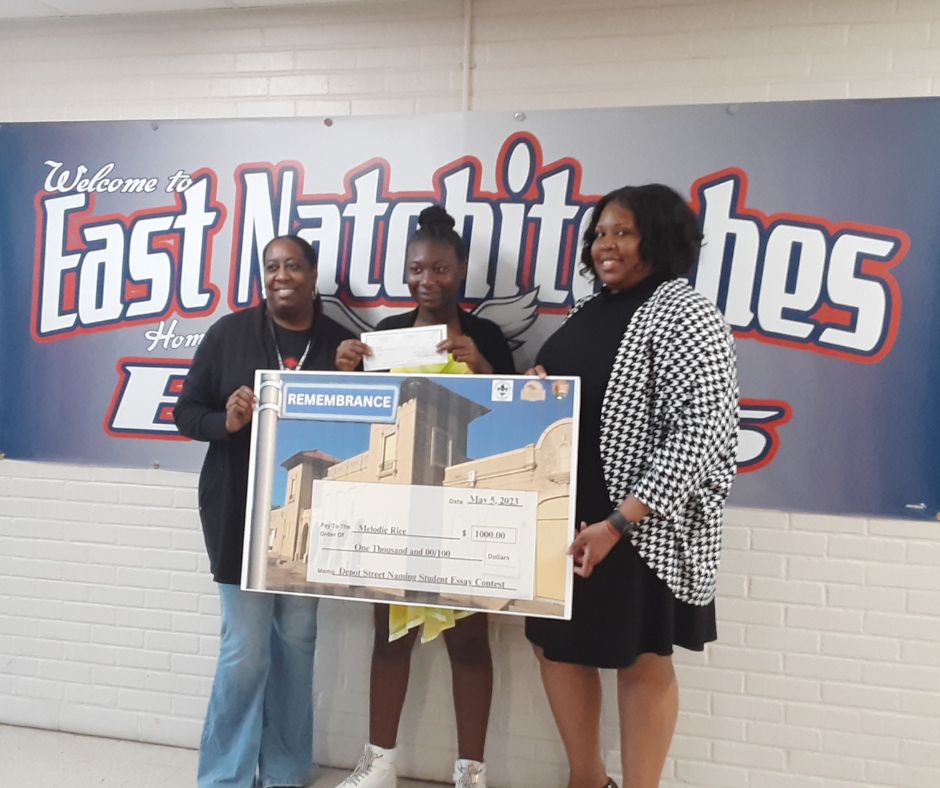 East Natchitoches 6th grader Melodie Rice wins student essay contest. Student holding large check stands between her teacher and principal in front of school sign.