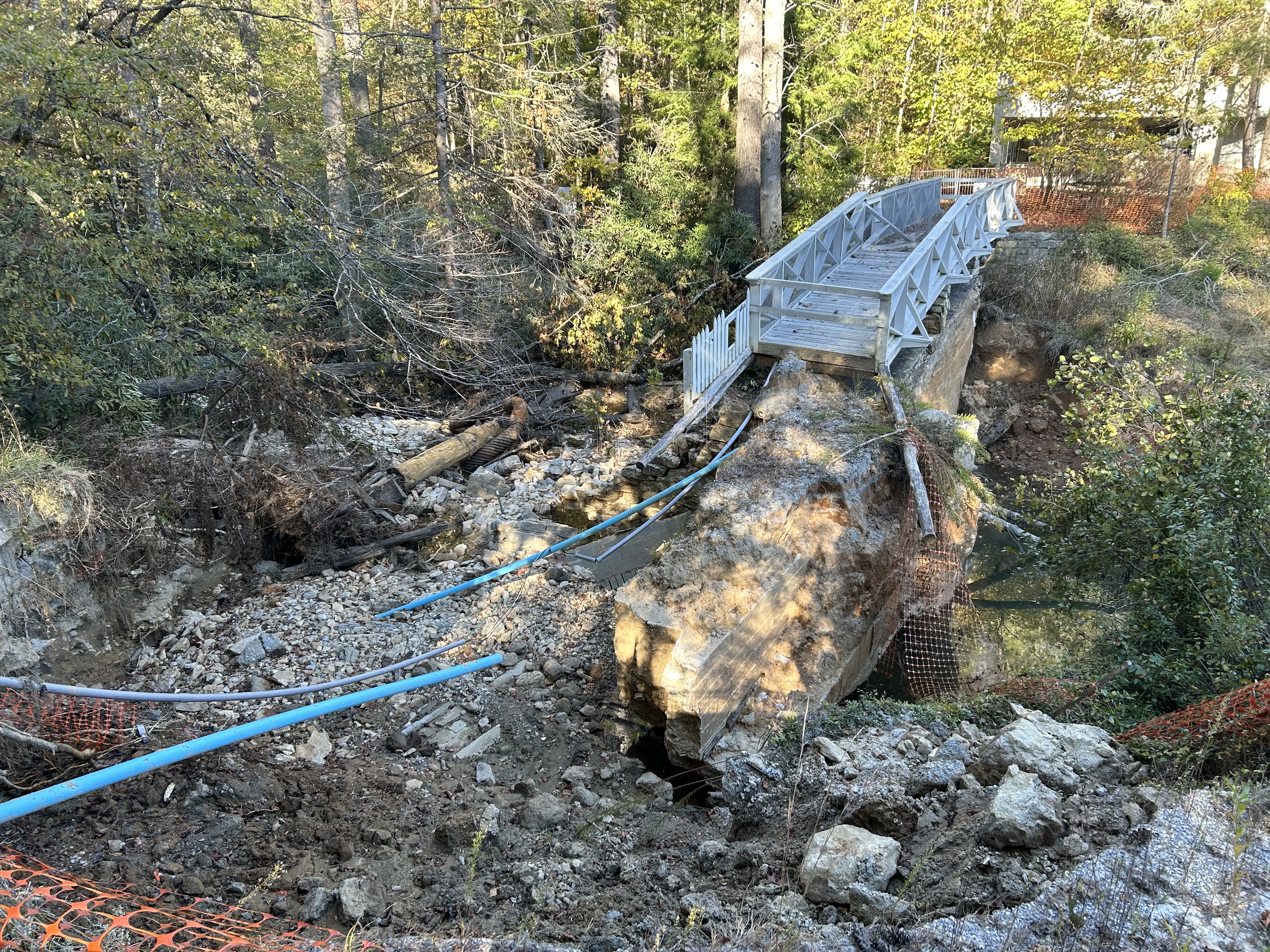 Front Lake Dam Damage A boardwalk and thick concrete wall end abruptly. They are surrounded by broken pipes and debris. Orange safety fencing is visible in the distance and foreground.