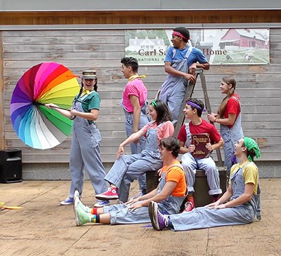 7 actors in colorful shirts and blue jean overalls act on a stage