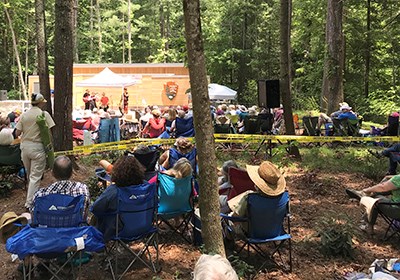 large crowd sits comfortably under the shade of trees while musicians play on a stage in the background