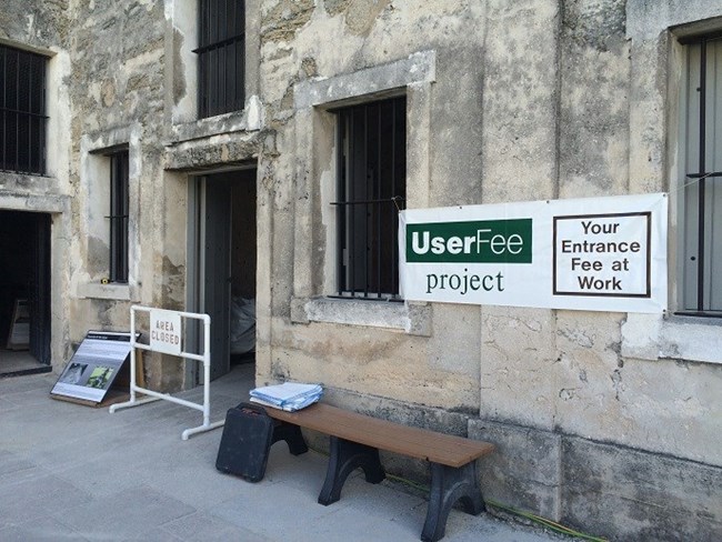 a banner stating "Use Fee Project, your entrance fee at work" hangs near the barricaded door to a room