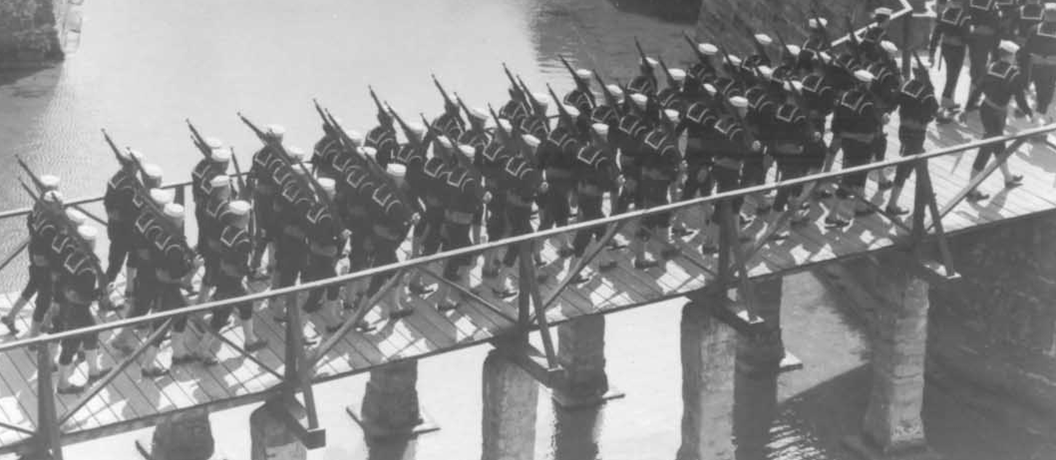 Coast Guardsmen march across the Castillo's drawbridge with shouldered weapons.