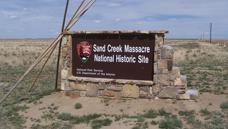 tipi poles lean against the entrance sign for Sand Creek Massacre National Historic Site