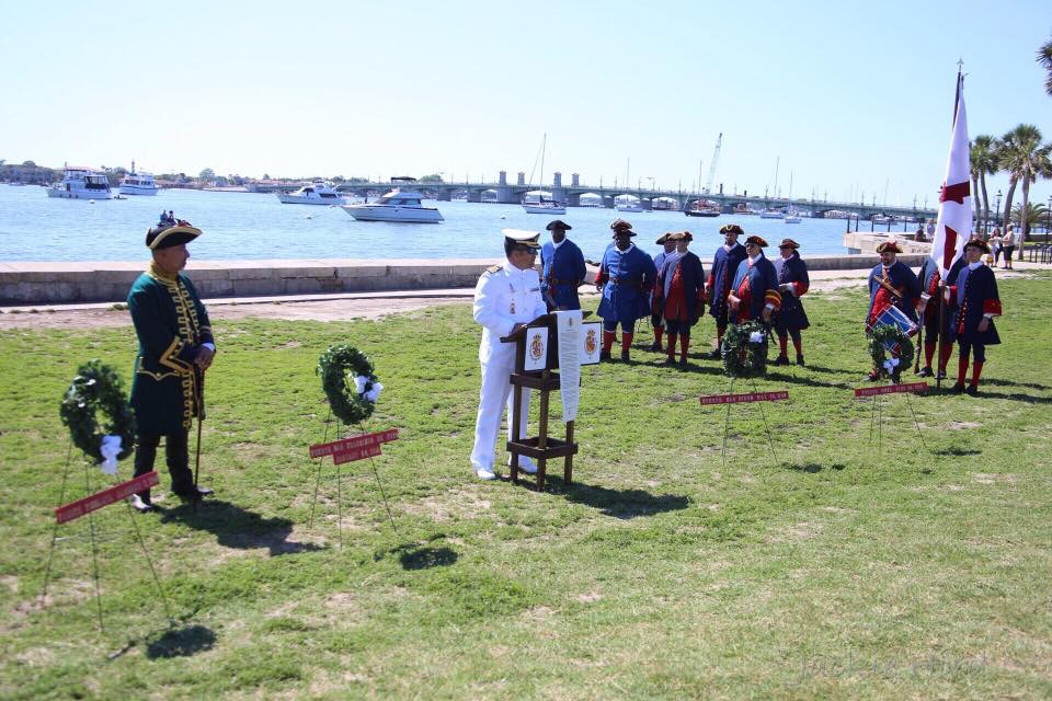 a man dressed in a military uniform gives a speech with living history spanish soldiers in the background