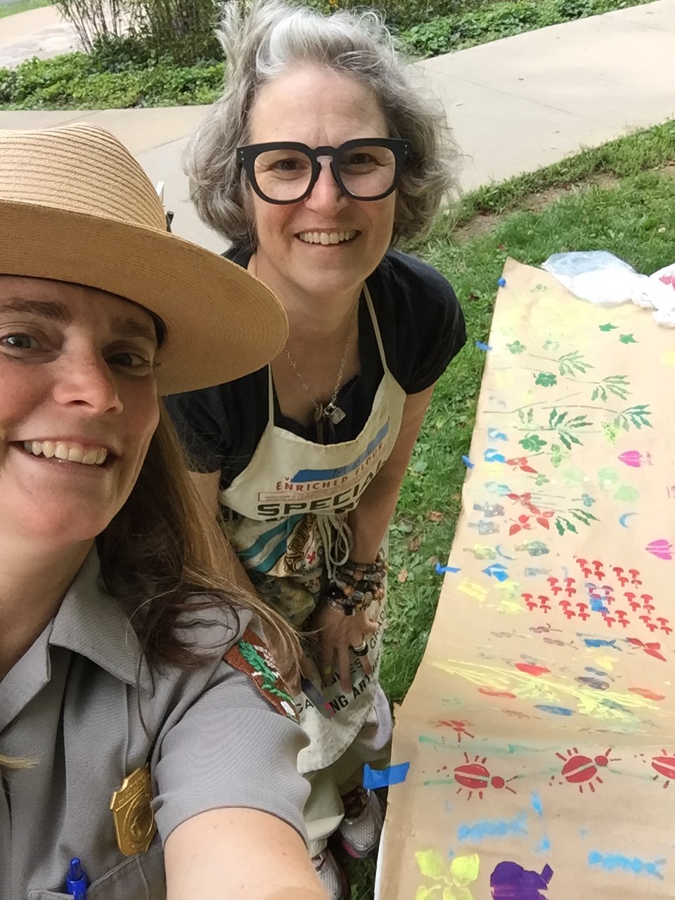 Two women, one in ranger uniform, behind a table covered in brown paper with multicolored stamps.