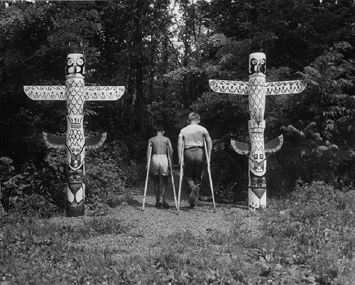 Two children using crutches face away from the camera. They are standing between two totem poles.