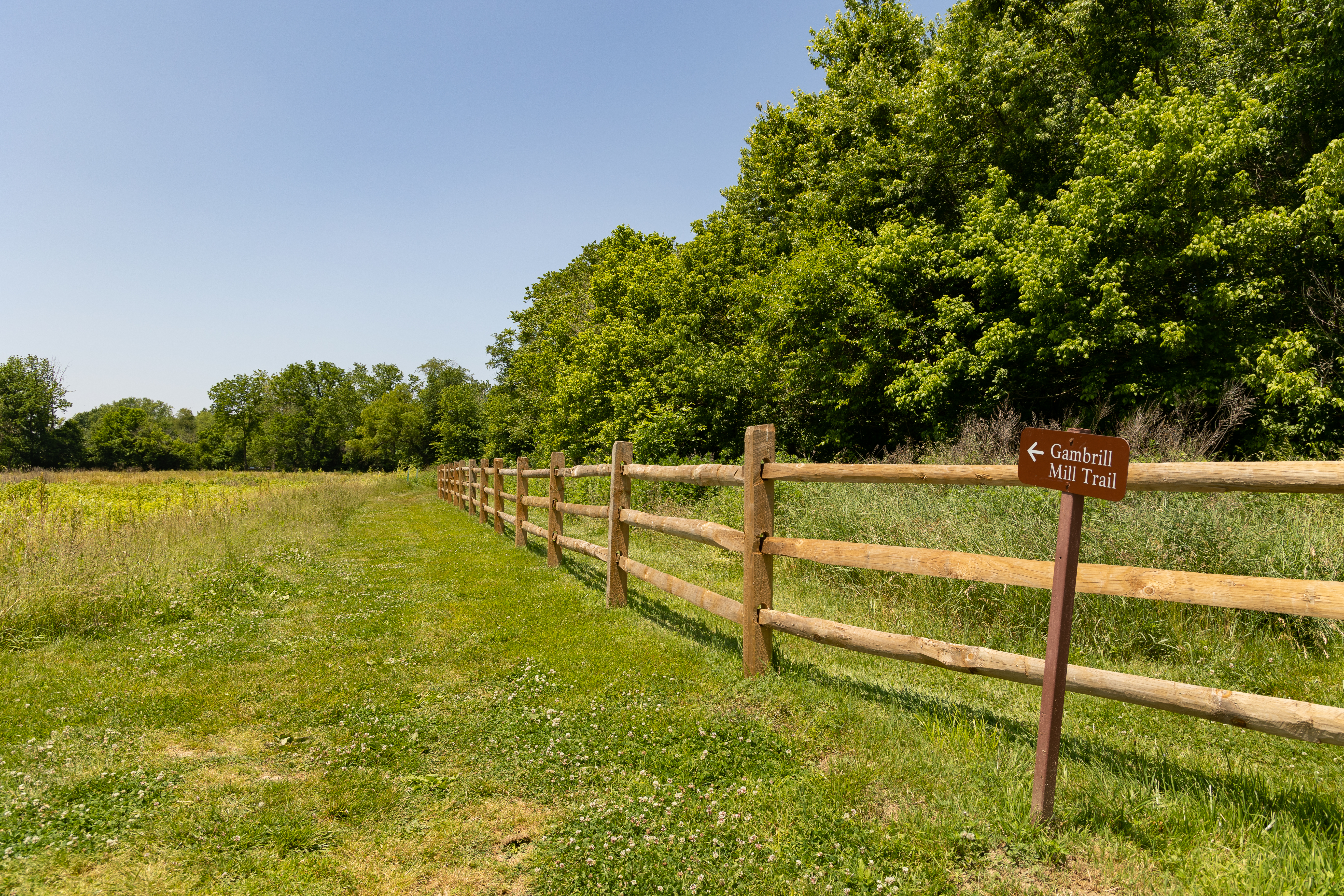 Gambrill Mill Trail An entrance to the Gambrill Mill Trail at Monocacy National Battlefield. The trail is a roughly six-foot-wide path of mowed grass. On the left side of the trail is a grassy field. On the right side of the trail is a wooden fence behind which is tall grass