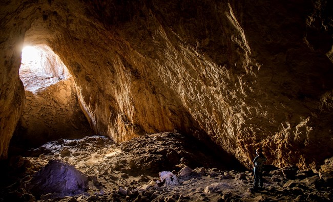 a caver stand in front of the entrance of goat cave. there is light shining through