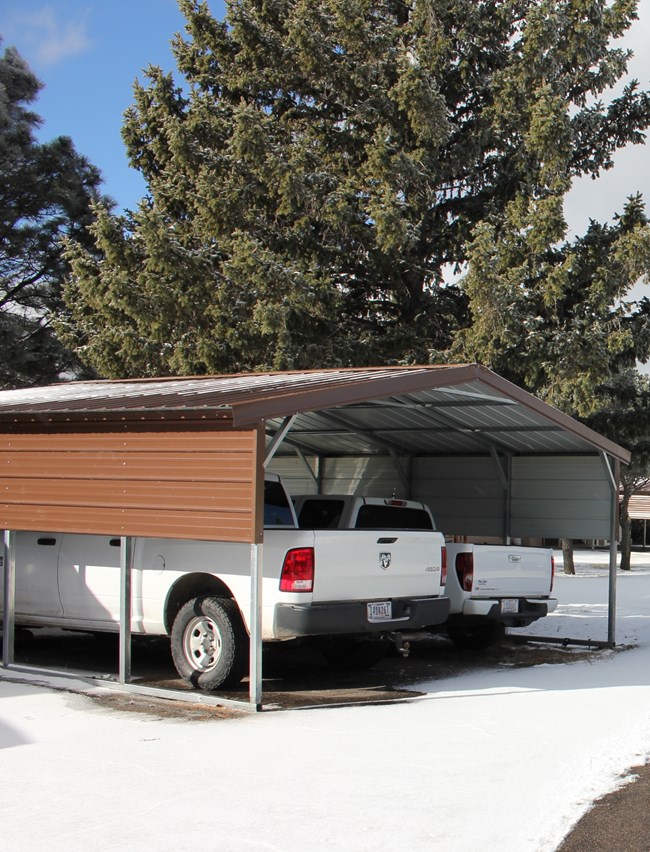 Two pickup trucks are parked inward underneath a carport.  Snow covers the ground while tall pine trees erupt into the blue sky.