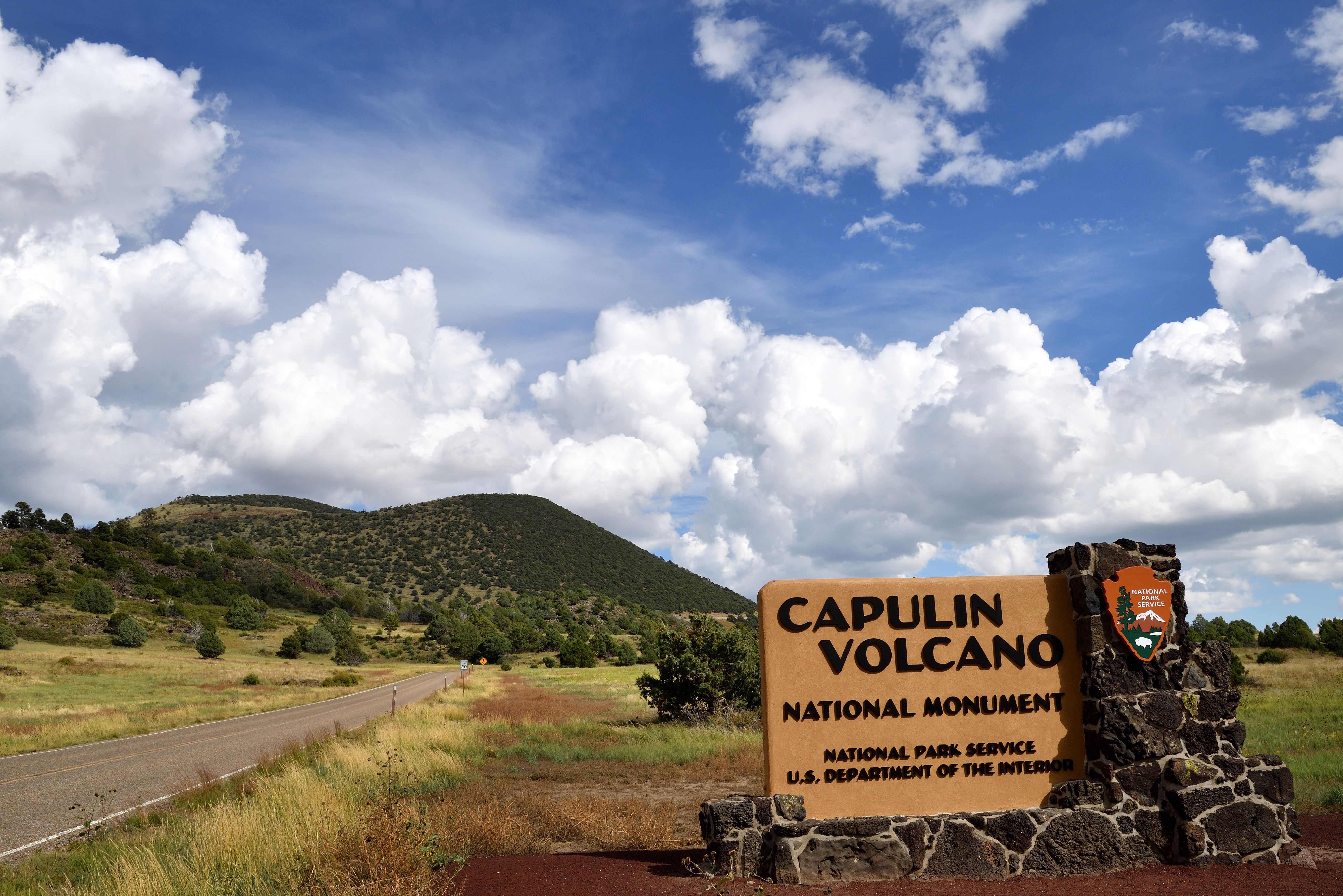An entrance sign reading "Capulin Volcano National Monument, National Park Service, U.S. Department of the Interior" is constructed of rough lava rock.  In the distance the volcano is dotted with trees and defines the horizon