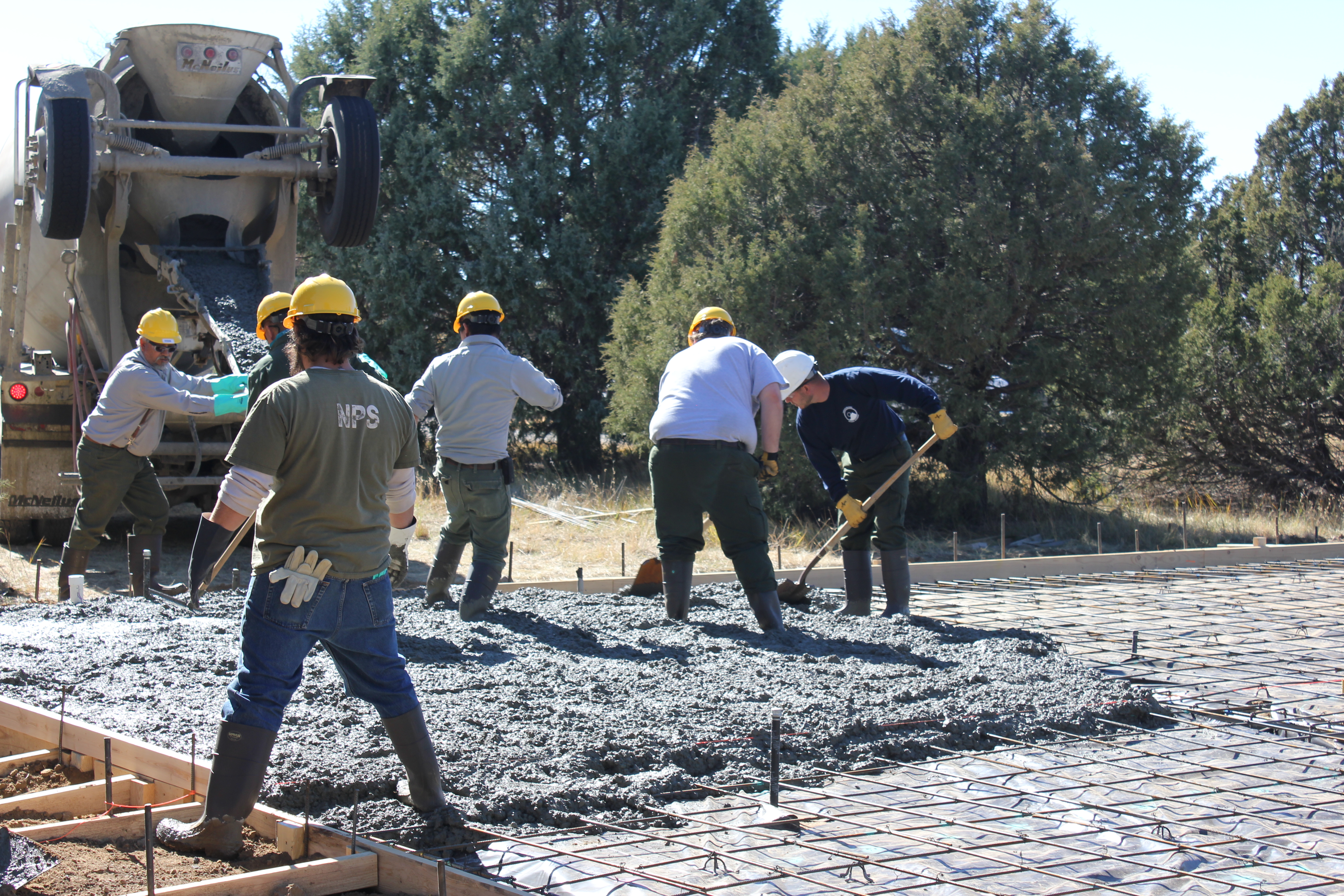 Workers stand in a pad of wet concrete while shoveling and smoothing it out.  A cement truck is backed up to the worksite.