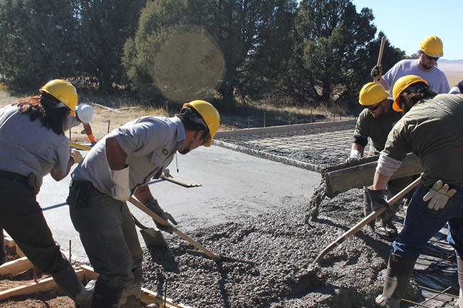 Five construction workers wear hardhats as they work to install a concrete foundation.  In the foreground workers shovel and smooth wet concrete which is dispensed from a trough to the right.  Trees line the close horizon.