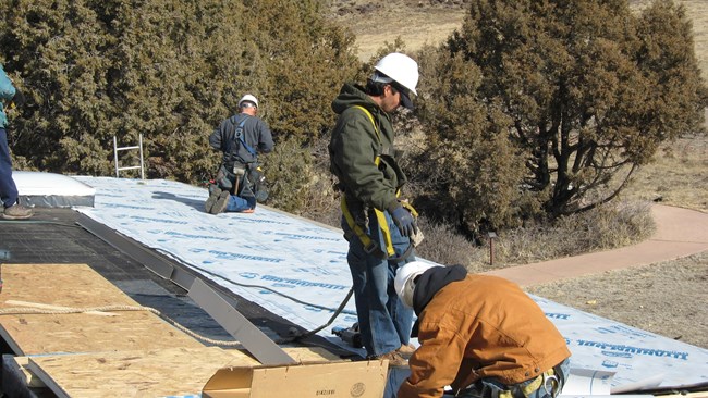 Four workers are repairing a flat roof.  The workers wear hard hats.  Two workers kneel while the other two are standing.  To the right down below a sidewalk disappears into the horizon which is full of trees.