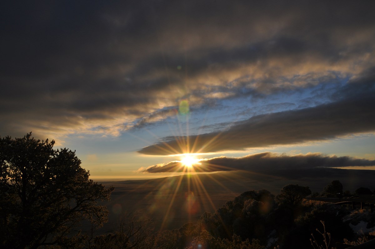 Sunrise 2 View from a high point onto the plains. The sun forces its way through clouds at the center of the picture where the rays appear as beams broadcasting out of the cloudy blue sky. Silhouetted trees are dappled around their edges by the soft glow of the sun