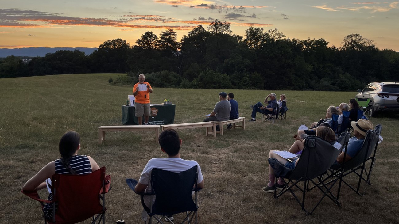 National Park Service volunteer talks to a seated crowd underneath a sunset sky.