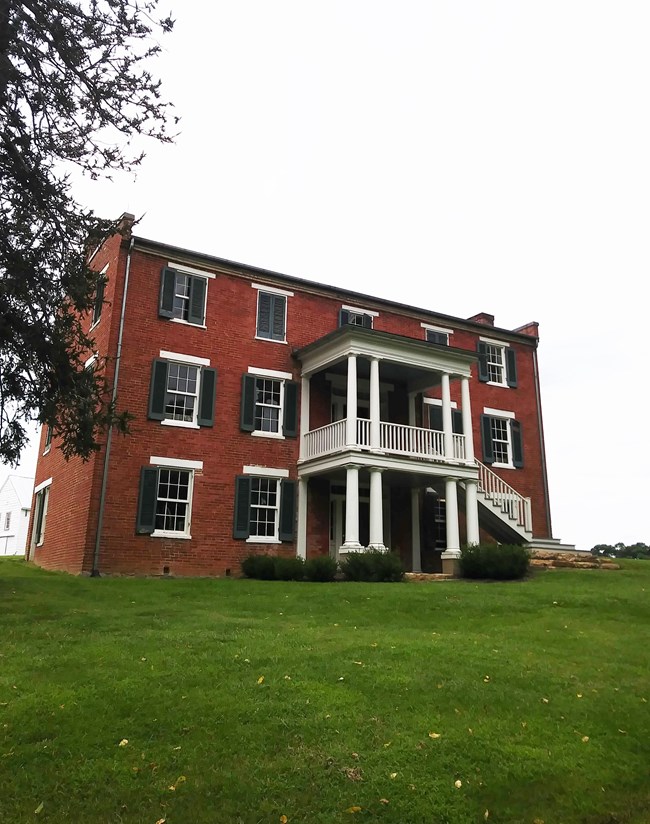 A brick three story Antebellum farmhouse sits atop a grassy hill.