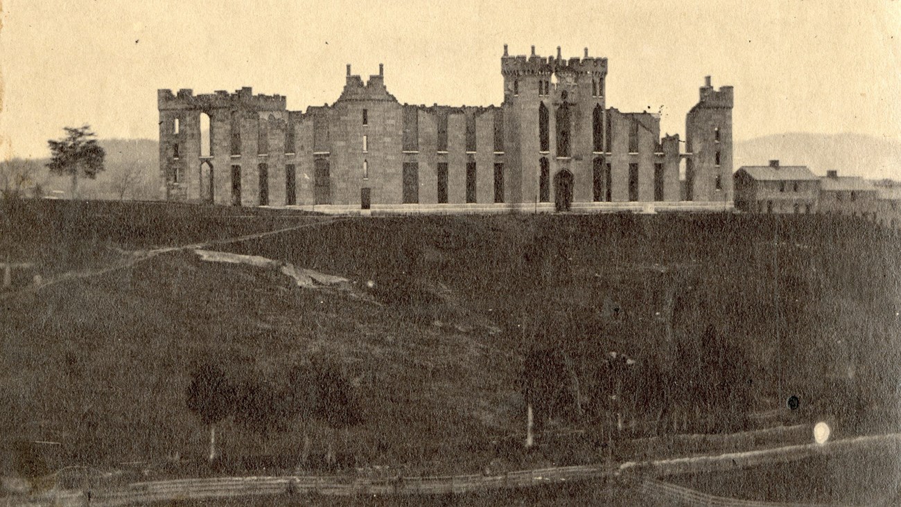 An 1800s photo show a burned out fortress-like school building atop a hill.