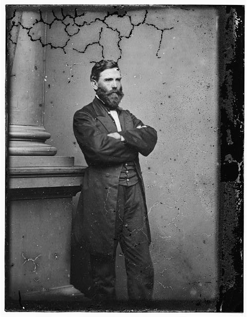 An 1860s studio photo shows a standing portrait of a bearded man, arms folded, wearing a long coat.
