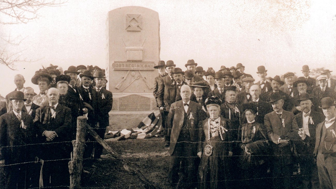 Well-dressed men and women pose in front of a stone war memorial in a 1907 photo.