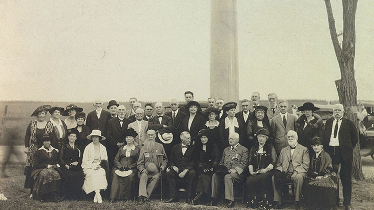 A 1920 photo captures well dressed men and women gathered in front of a stone column war memorial.