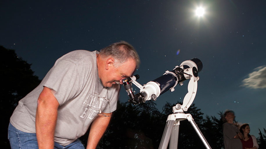 A man looks at stars through a refracting telescope in a field.