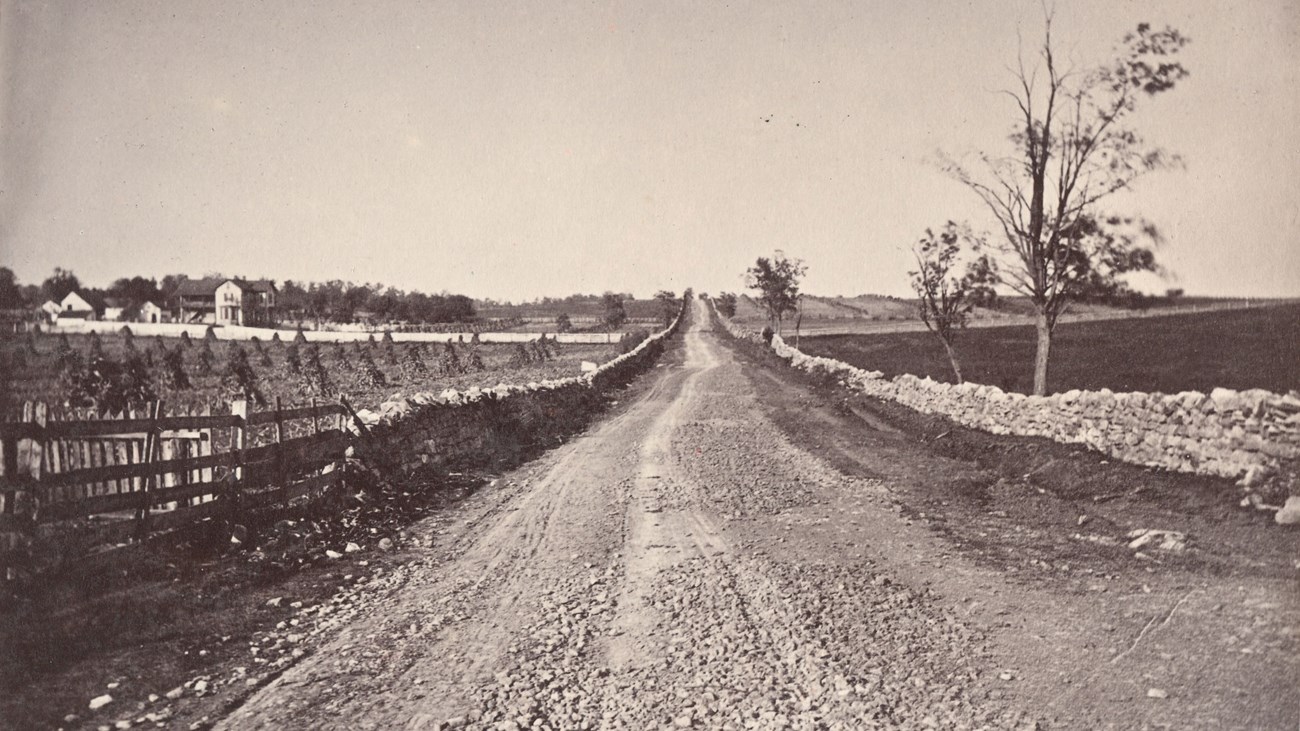 An 1800s photo shows a stone wall lined country road through farmland.