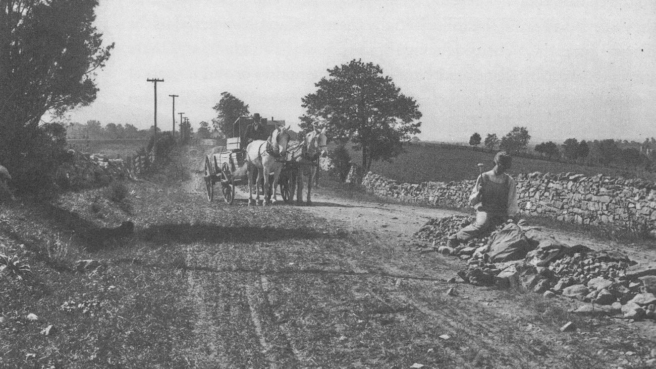 An early 1900s photo shows a workman breaking rocks on a country road with a horse drawn carriage behind him.
