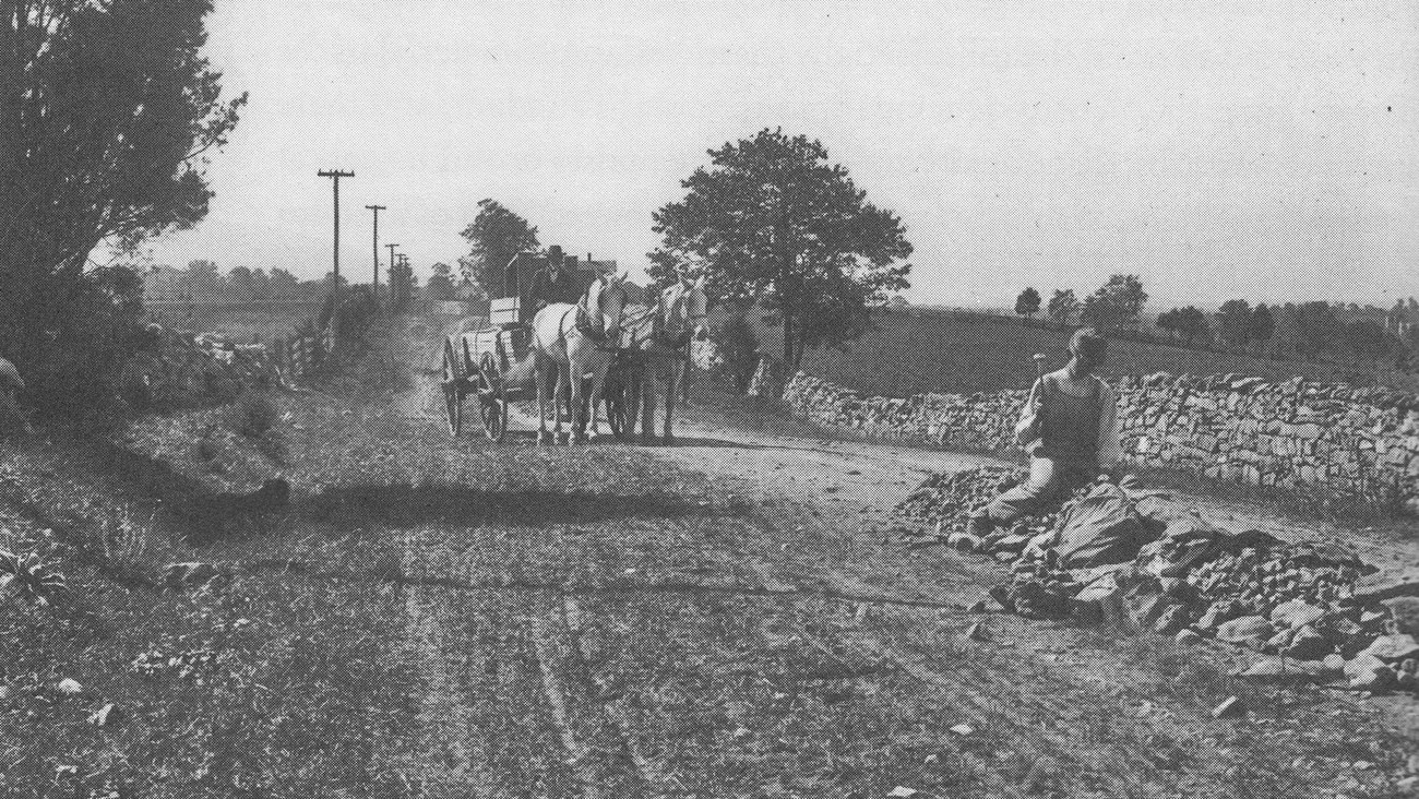An early 1900s photo shows a workman breaking rocks on a country road with a horse drawn carriage behind him.