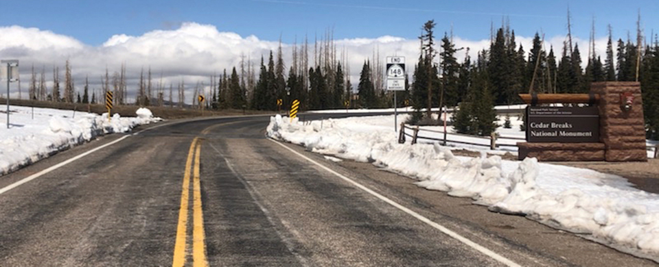 A two lane road leads passed a park sign, snow covers the ground on either side.