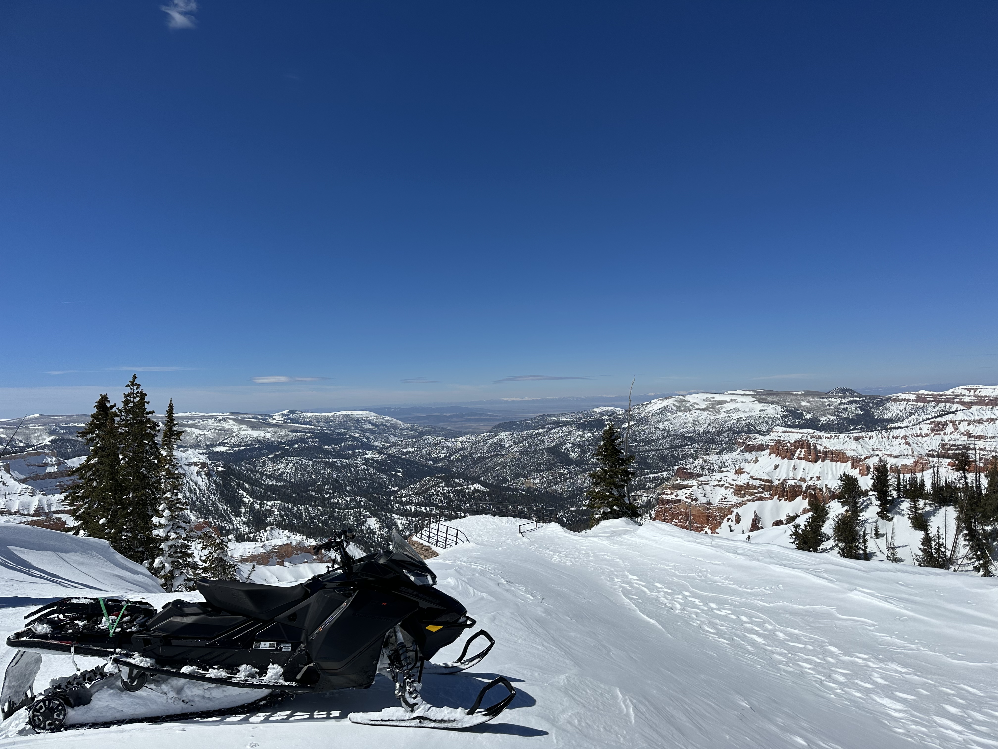 A Snowmobile is parked on the snow covered park road, near the Sunset View Overlook, looking west over a snowy landscape and cloudless sky.