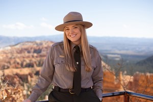 NPS ranger Allana Olbrich smiles for a portrait while standing at a railing overlooking the Cedar Breaks amphitheater. She wears a ranger uniform and wide-brimmed hat.