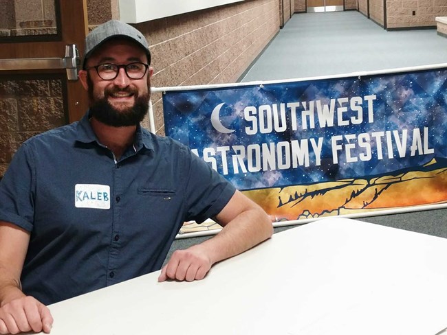 Southwest Astronomy Festival Volunteer Man with beard sitting at desk with sign saying "SW astronomy festival" in background.