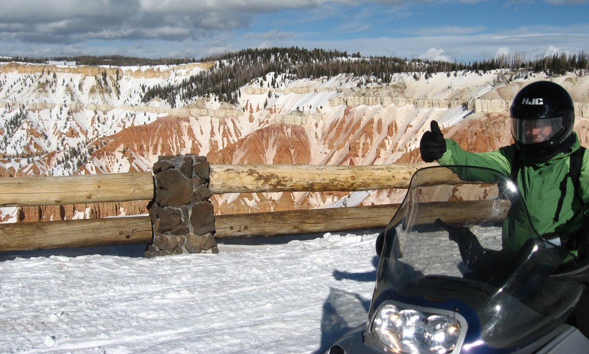 A snowmobiler gives a thumbs up in front of a scenic overlook.