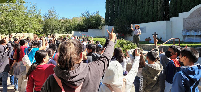 A park ranger stands in front of a group of school children in an outdoor garden holding two fingers in the air while the children do the same