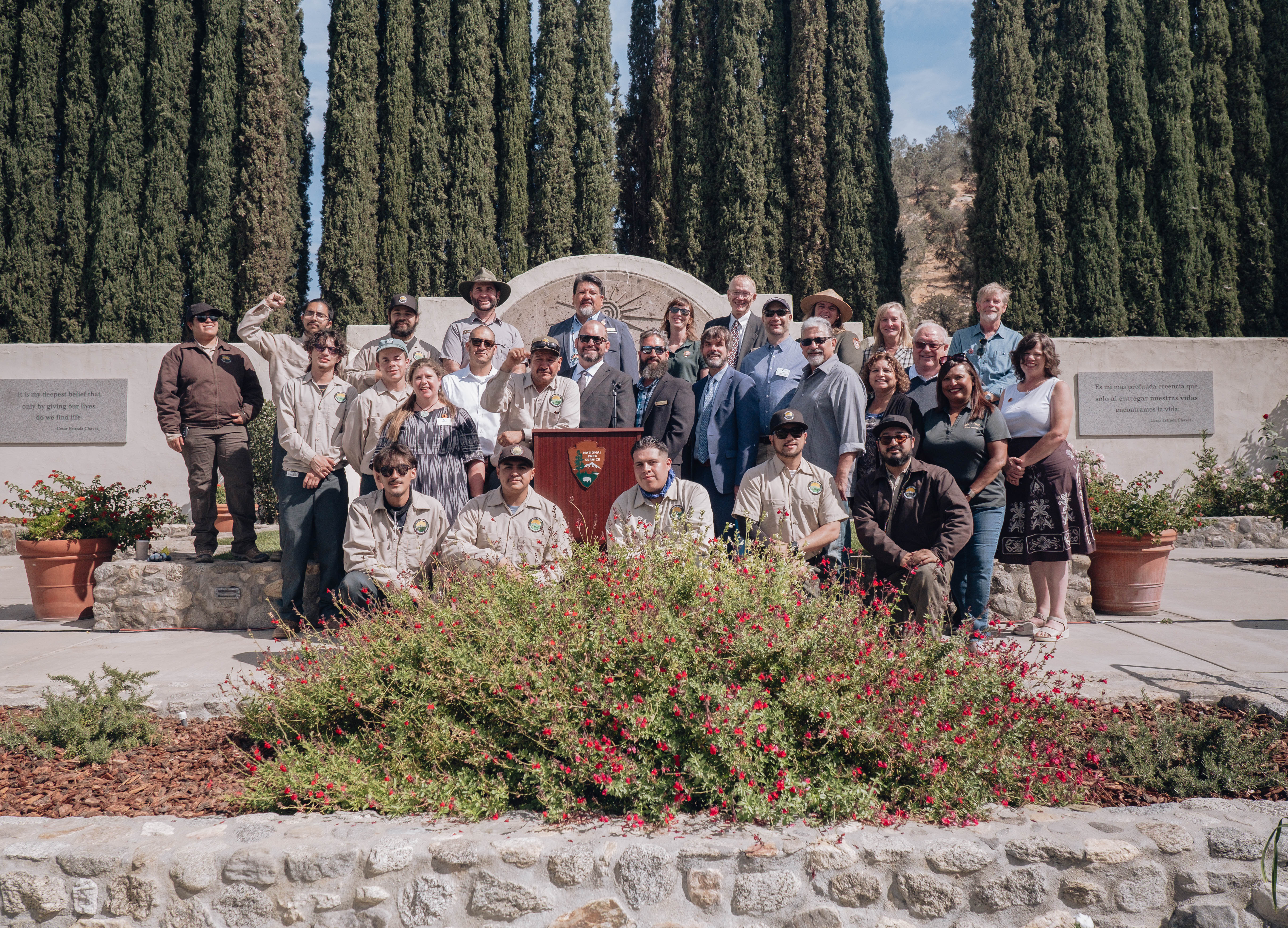 Group photo taken in the memorial Garden Three rows of people arranged around a podium with large plant with red flowers in the foreground and tall narrow trees in background
