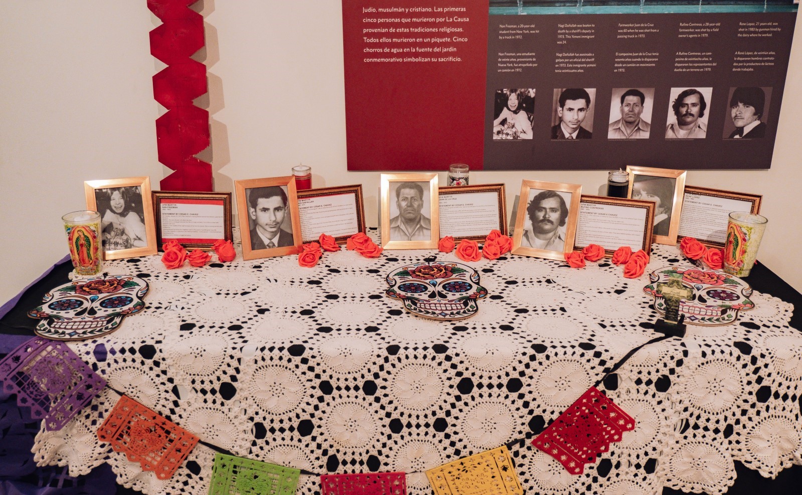Dia de los Muertos altar honoring the five martyrs of the farmworker movement A set of 5 framed photographs next to framed sheets of text sit on a black table covered with a white lace tablecloth. Simliar photos are on the wall. Also on the table are 3 colorful mexican-style day of the dead skulls, 5 candles in tall glass holders.