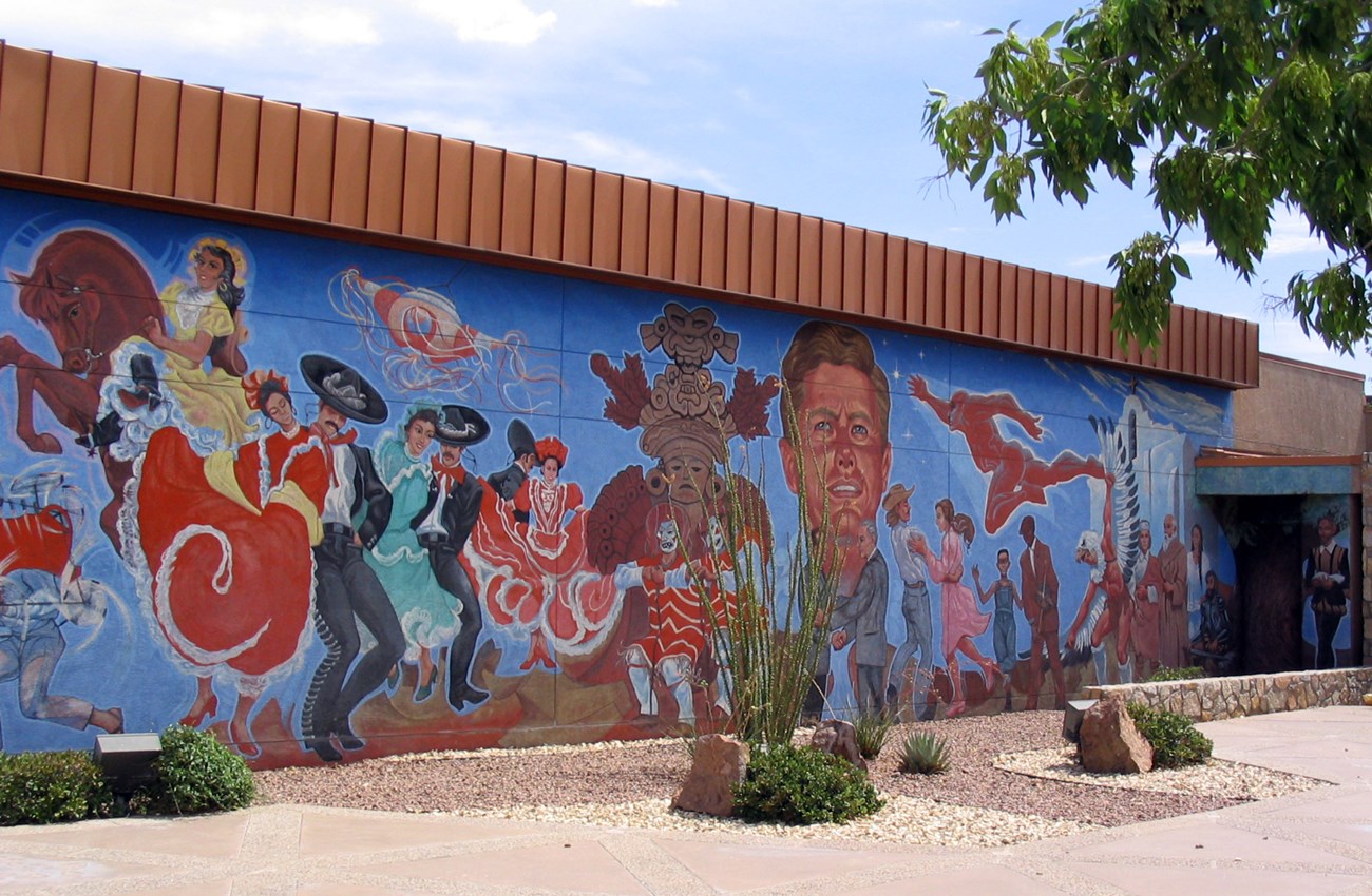 Long mural panel with cultural images from Mexico, United States, and Spain. Large head of President Kennedy is in the middle with Presidents Johnson and Lopez Mateos shaking hands below.